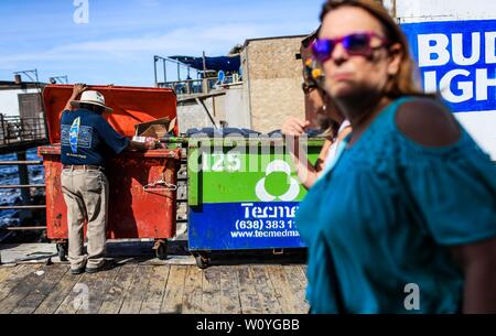 Puerto Peñasco, Sonora, October 20, 2017.- Two American women tourists stroll the tourist dock of Puerto Peñasco while a May adult man of extreme poverty looks for food inside the trash contents. Dos mujeres turistas norteamericanas pasean por el muelle turístico de Puerto Peñasco mientras un hombre adulto mayo de pobreza extrema  busca comida dentro de los contenidos de basura. (Foto: Luis Gutierrez /NortePhoto.com) Stock Photo