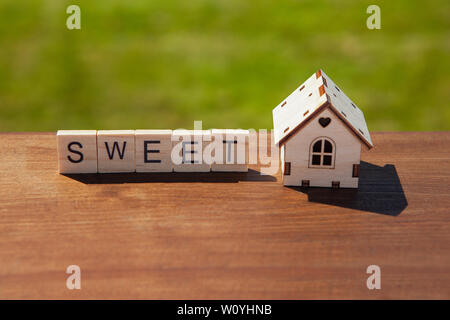 Word sweet of wooden letters and small toy wooden house on brown surface, green grass in background. Concept sweet home, mortgage, dream house Stock Photo