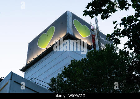 Grenfell Tower at dusk, now covered in tarpaulin, with the symbolic green heart displayed. 'Forever in our hearts' Stock Photo