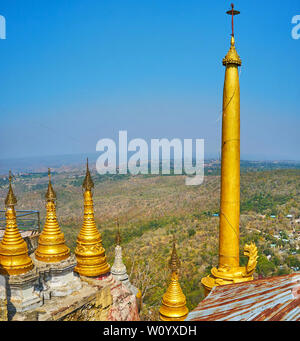 The small golden stupas and tall slender Buddhist pillar in Taung Kalat monastery  on the edge of Popa mountain outcrop, with a view on green hills og Stock Photo