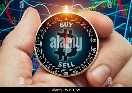 hand holding compass with glowing bitcoin symbol and in front of stock market chart data. Compass needle showing buy word. Stock Photo