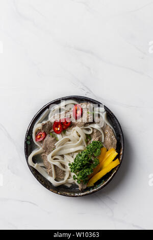 Hot noodle soup with beef, egg, mushrooms and vegetables in black bowl on white marble background. Copy space for text. Stock Photo