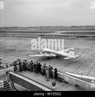 A Vintage 1950s black and white photograph taken at London Heathrow Airport, showing a group of people standing and looking over a balcony towards a Flugfelag (Iceland Airways), Douglas C-54 (DC-4) airliner. Stock Photo