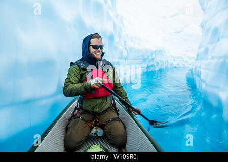 Man in harness with ice climbing gear and pfd paddling an inflatable canoe through a crevasse filled with deep blue water from the melting glacier. Stock Photo