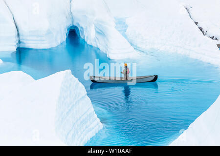 Man paddles a canoe in front of a small flooded ice cave on a glacier lake in the Alaskan wilderness. Stock Photo