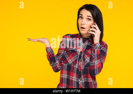 Amazed woman keeping mouth wide open, looking surprised, talking on mobile phone, conducting pleasant conversation isolated on yellow background. Peop Stock Photo