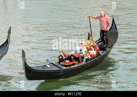 Two tourists sitting in the gondola with the gondolier, typical Venetian row boat. Sightseeing tour along the Grand Canal of the famous city Stock Photo