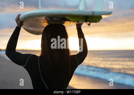 Back view of a young woman in the beach holding on her surfboard