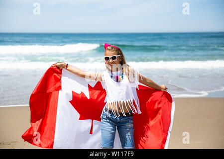 Happy Canadian girl carries fluttering white red flag of Canada against blue sky and ocean background. Canadian flag is a symbol of freedom, liberty Stock Photo