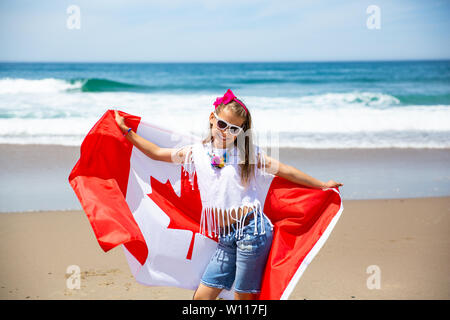 Happy Canadian girl carries fluttering white red flag of Canada against blue sky and ocean background. Canadian flag is a symbol of freedom, liberty Stock Photo