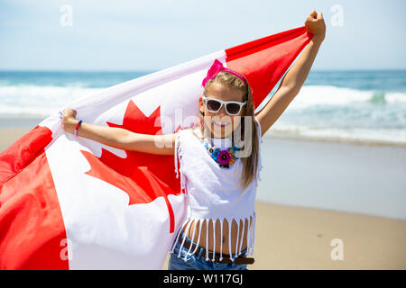 Happy Canadian girl carries fluttering white red flag of Canada against blue sky and ocean background. Canadian flag is a symbol of freedom, liberty Stock Photo