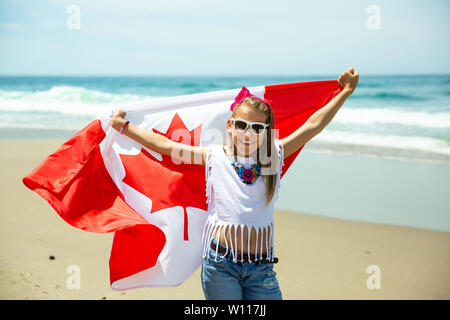 Happy Canadian girl carries fluttering white red flag of Canada against blue sky and ocean background. Canadian flag is a symbol of freedom, liberty Stock Photo