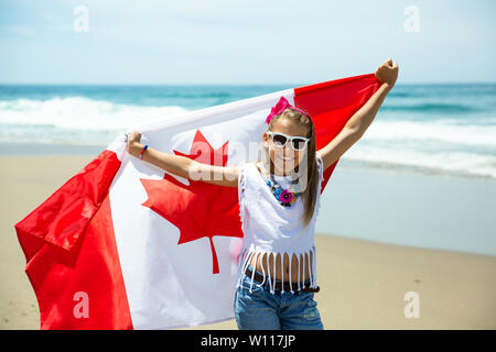 Happy Canadian girl carries fluttering white red flag of Canada against blue sky and ocean background. Canadian flag is a symbol of freedom, liberty Stock Photo