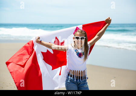 Happy Canadian girl carries fluttering white red flag of Canada against blue sky and ocean background. Canadian flag is a symbol of freedom, liberty Stock Photo