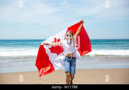 Happy Canadian girl carries fluttering white red flag of Canada against blue sky and ocean background. Canadian flag is a symbol of freedom, liberty Stock Photo