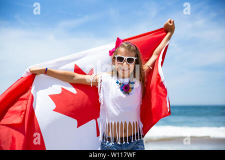 Happy Canadian girl carries fluttering white red flag of Canada against blue sky and ocean background. Canadian flag is a symbol of freedom, liberty Stock Photo