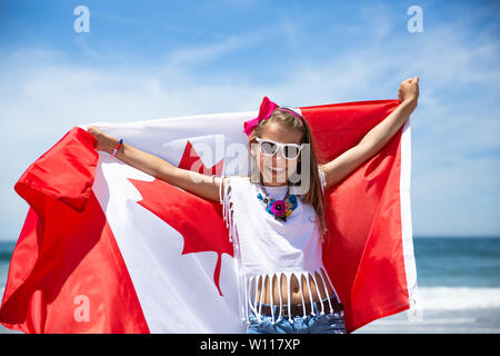 Happy Canadian girl carries fluttering white red flag of Canada against blue sky and ocean background. Canadian flag is a symbol of freedom, liberty Stock Photo