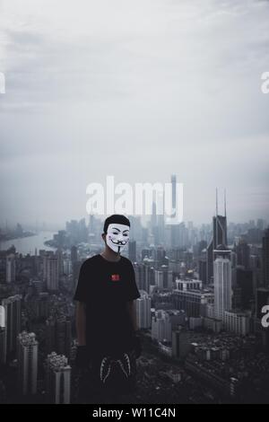 Male wearing an anonymous mask standing on a roof with an amazing urban city in the back Stock Photo