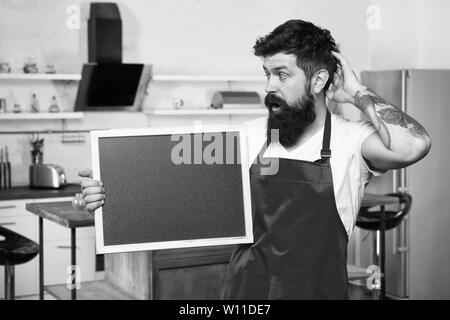 What to cook. urprise. This is what you need. Hipster in kitchen. Restaurant or cafe menu. Advertisement. Bearded man in red apron. Mature male. Bearded man cook. Man chef with board, copy space. Stock Photo