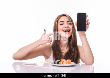 Smiling young asian woman eating sushi at the table isolated over white background, showing blank screen mobile phone Stock Photo