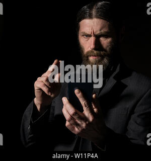 Old believer senior priest with a smartphone, bearded old man is calling on a dark background. Stock Photo