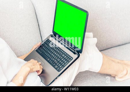 Middle-aged woman on the gray sofa working on green screen laptop, soft focus Stock Photo