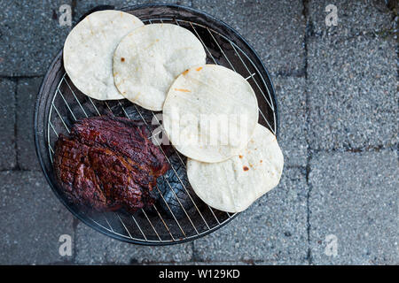 A large beef brisket and and corn tortillas being barbequed on a small charcoal hibachi grill with glowing coals underneath. Stock Photo
