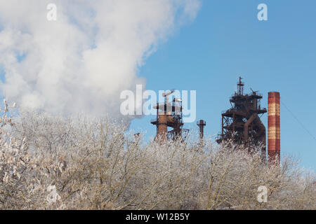 metallurgical plant with cloud of pollution exhaust with frozen winter bush in foreground at daylight. Stock Photo