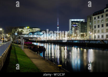 Illuminated Friedrichstrasse train station and other buildings by the Spree River and Fernsehturm TV Tower in Berlin, Germany, at dusk. Stock Photo