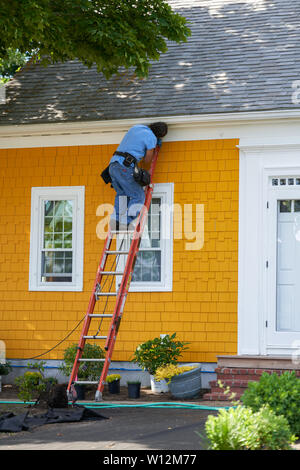 Wellfleet, MA - June 12, 2019: Man is painting trim on a bright yellow house as part of a renovation project including new plantings at the base of th Stock Photo