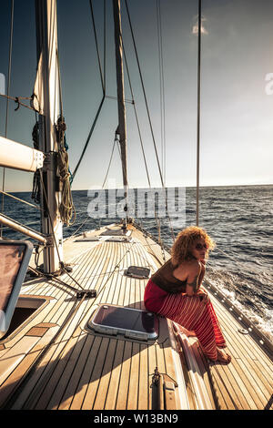 Travel people influencer social media concept with cute curly young adult woman sit down on a sail boat in the ocean enjoying the sun and the freedom Stock Photo