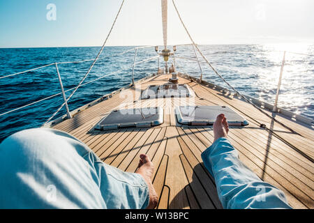 Happiness and relax concept for traveler people - man legs point of view on a wooden sail boat with sun and blue ocean around - luxury and holiday vac Stock Photo