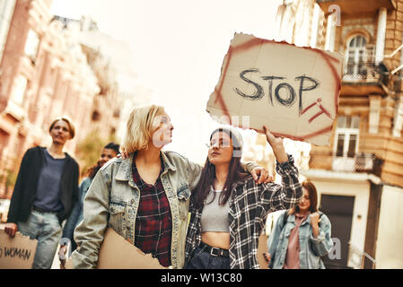 Make peace Young woman in eyewear and casual clothing is holding a banner with word stop it while standing on the road during a women's march aroud female activists. Human rights. Protest concept Stock Photo