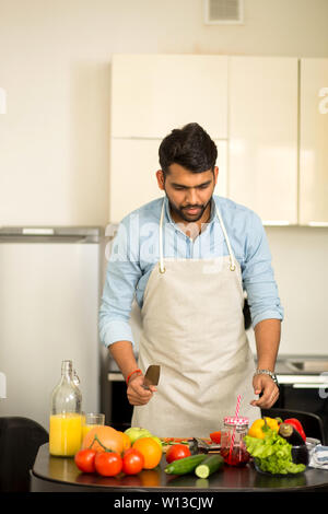 Attractive bearded indian man in blue shirt and apron preparing salad with tomatoes, cucumber, sweet pepper, lettuce in the kitchen. Healthy and veget Stock Photo