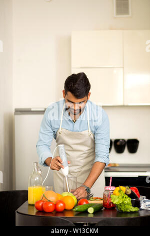 Young indian bearded male wearing shirt and apron cooking fresh vitamin juice, smoothie, cocktail with raw vegetables and fruits in blender in kitchen Stock Photo