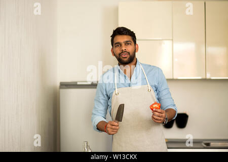 Handsome bearded amazed indian man, preparing salad in kitchen, holding knife and tomato looking at camera, cooking in kitchen at home. Healthy, veget Stock Photo