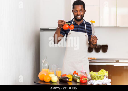 Joyful charming young black man in apron preparing organic vegetable salad and fruit juice in kitchen at home, holding in hands ripe tomatoes, knife a Stock Photo
