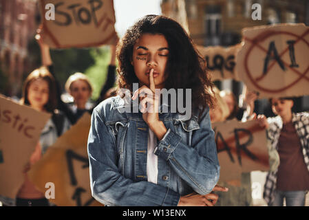 Young afro american woman with word power written on her face and with closed eyes showing silence sign while protesting with group of female activists outdoors on road. Human rights. Demonstration. Women power Stock Photo