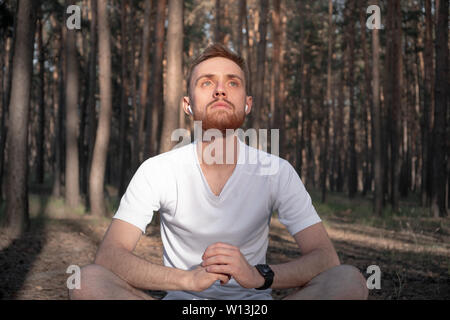 Young male person meditating in the forest using modern technology. Active man sits in the pine woods and enjoys the meditation outdoors Stock Photo