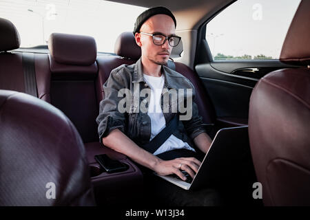 Young business person using laptop on a backseat of a car. Milennial person using modern technology while commuting by a private or rental car Stock Photo
