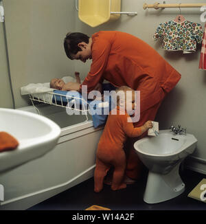 1960s mother. A young woman is cleaning and changing diapers on her baby that lies on a changing table. An older child is standing beside hugging her to get attention. The clothes and colors are typical 1960s. Sweden 1964 Stock Photo