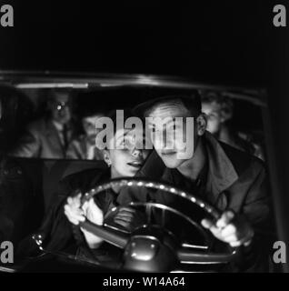 Driving in the 1950s. A young couple in the driving seat. He is focused on driving in the dark night and his eyes are on the road. She is more interested in him and sits as close to him as she can. Sweden 1959. ref BV100-2 Stock Photo