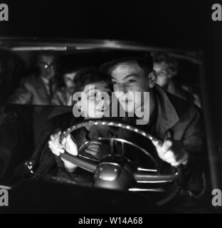 Driving in the 1950s. A young couple in the driving seat. He is focused on driving in the dark night and his eyes are on the road. She is more interested in him and sits as close to him as she can. Sweden 1959. ref BV105-5 Stock Photo