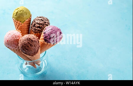 Various ice cream scoops in cones with copy space. Colorful ice cream in cones chocolate, strawberry, blueberry, pistachio or matcha, biscuits chocolate sandwich cookies on blue background. Banner Stock Photo