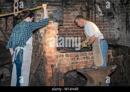 A male blacksmith working at a forge with a female pumping the bellows. Stock Photo