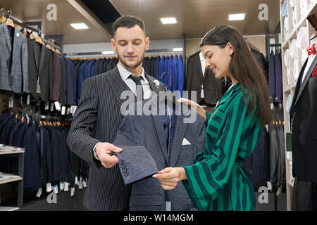 Handsome client choosing suit in fashionable boutigue. Pretty girl, shop assistant holding jacket on hanger, showing and proposing. Man and shop consu Stock Photo