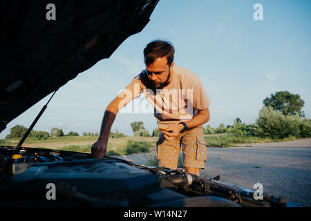 The guy in the pink t-shirt checks the engine of a black car lifting the hood in the country outside the road Stock Photo
