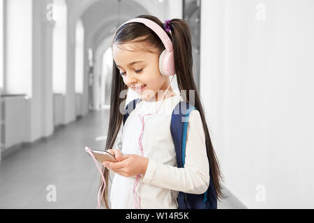 Happy school girl using smartphone and listening music from pink headphones on break. Pretty pupil having fun, watching videos at call phone. Free time of lessons in school. Stock Photo