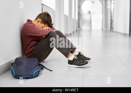 Side view of sad school boy sitting alone on floor at corridor and hiding face by hands. Brunet boy having bad mark and terrible mood after lessons, crying. Hard period of teenagers and studying. Stock Photo