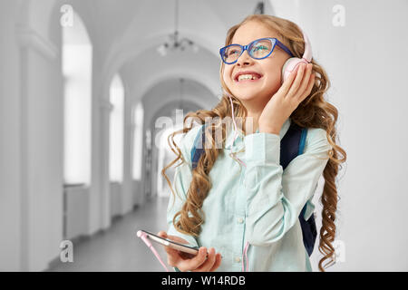 Smiling pretty school girl wearing eyeglasses and backpack listening music from big pink headphones, looking away and dreaming. Funny girl resting after lessons, using smartphone and going home. Stock Photo